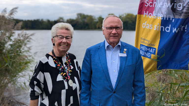 Zwei Personen vor See und Beachflag mit Aufschrift "Schleswig-Holstein, das sind wir!"