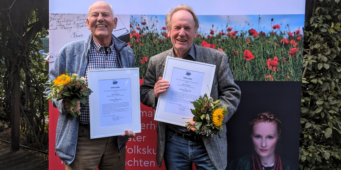 2 Männer mit gerahmter Urkunde vor Messewand