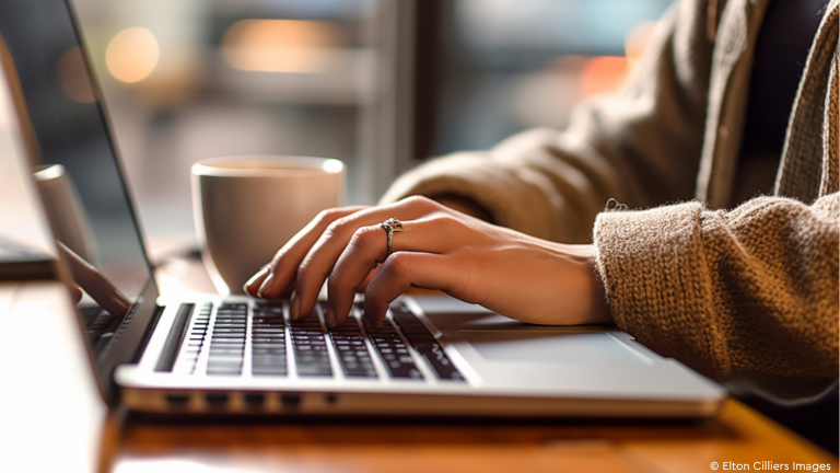 Frauenhand auf Laptoptastatur mit Kaffeetasse im Hintergrund